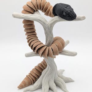 May include: A segmented, articulated snake figure with a black head and a tan, ribbed body. The snake is coiled around a light grey, textured tree branch stand. The figure features a detailed scale pattern on the head and a flexible, multi-piece construction that allows for movement along the length of the body.
