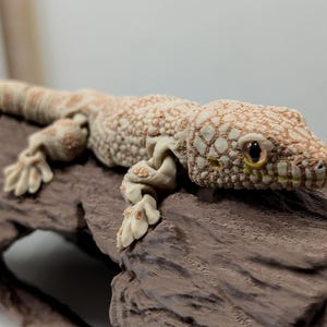 以下が含まれることがあります： ベージュとライトブラウンの関節式ヤモリのフィギュアで、表面は凹凸のある質感で、手足に関節が見えます。トカゲは、木目調の仕上げが施された、暗い茶色の中空で質感のある丸太のような構造物の上に配置されています。