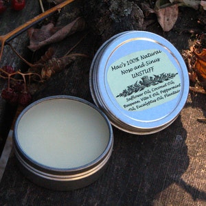 May include: A silver tin with a green label that reads "Mac's 100% Natural Nose and Sinus Unstuff" and lists the ingredients: Safflower Oil, Coconut Oil, Beeswax, Vita E Oil, Peppermint Oil, Eucalyptus Oil, Plantair. The tin is open and the inside is filled with a light-colored balm.