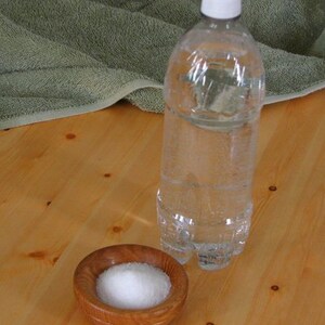May include: A clear plastic bottle of water with a white cap, sitting on a wooden surface next to a small wooden bowl filled with white salt.