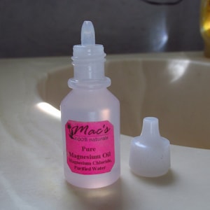 May include: A clear plastic bottle with a pink label that reads "Mac's 100% natural Pure Magnesium Oil Magnesium Chloride Purified Water". The bottle has a white dropper top and is sitting on a white surface.