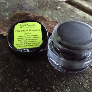 May include: A small glass jar with a black lid containing a black salve. The lid has a yellow label with the text "Mac's 100% Natural Old Black Drawing Salve" and a list of ingredients including sunflower oil, plantain, burdock, yarrow, comfrey, tea tree, frankincense, pine sap, lavender, beeswax, whiskey, activated charcoal, and bentonite clay.