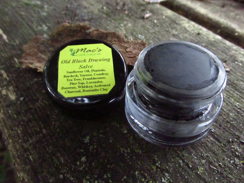 May include: A small glass jar with a black lid containing a black salve. The lid has a yellow label with the text "Mac's 100% Natural Old Black Drawing Salve" and a list of ingredients including sunflower oil, plantain, burdock, yarrow, comfrey, tea tree, frankincense, pine sap, lavender, beeswax, whiskey, activated charcoal, and bentonite clay.