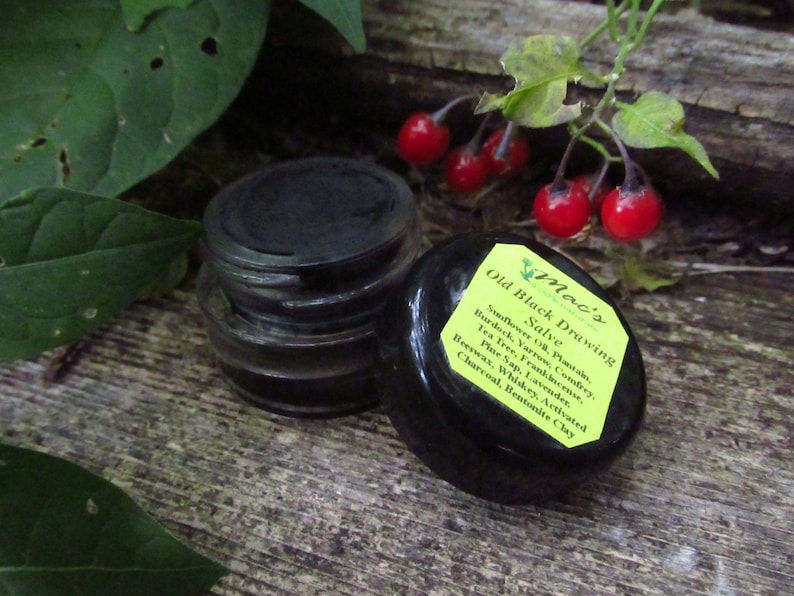 May include: A black jar with a black lid labeled "Mac's Old Black Drawing Salve". The label lists ingredients including sunflower oil, plantain, burdock, yarrow, comfrey, tea tree, frankincense, pine sap, lavender, beeswax, whiskey, activated charcoal, and bentonite clay.