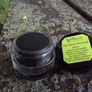 May include: A small black jar with a black lid labeled "Mac's Old Black Drawing Salve" with a list of ingredients including sunflower oil, plantain, burdock, yarrow, comfrey, tea tree, frankincense, pine sap, lavender, beeswax, whiskey, activated charcoal, and bentonite clay.