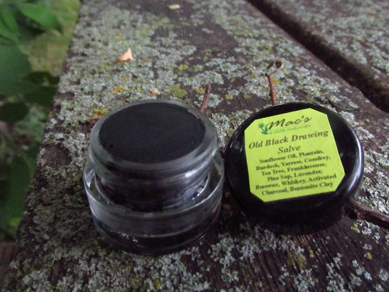 May include: A small black jar with a black lid labeled "Mac's Old Black Drawing Salve" with a list of ingredients including sunflower oil, plantain, burdock, yarrow, comfrey, tea tree, frankincense, pine sap, lavender, beeswax, whiskey, activated charcoal, and bentonite clay.