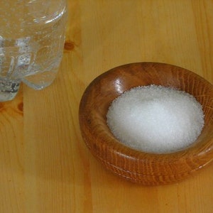 May include: A small wooden bowl filled with white granulated sugar. The bowl is sitting on a light wood surface.
