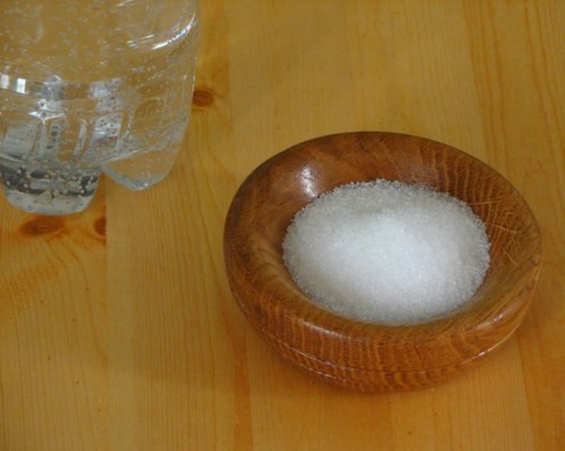 May include: A small wooden bowl filled with white granulated sugar. The bowl is sitting on a light wood surface.