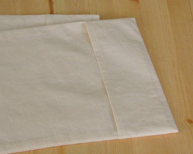 May include: Close-up of two off-white pillowcases, folded and arranged on a light wooden surface. The fabric appears to be a soft, plain weave. The pillowcases are neatly folded, showcasing the clean lines and simple design.