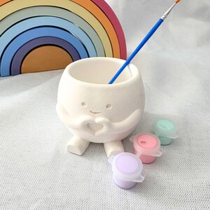 May include: A white ceramic planter shaped like a round character with small feet and arms forming a heart shape on its chest. A blue paintbrush rests inside the opening, accompanied by three small paint pots in pastel purple, pink, and green.