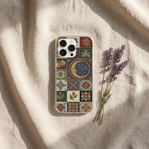 May include: A clear smartphone case featuring a grid of square tiles with various illustrations including a crescent moon, a bee, flowers, and fruit in shades of red, blue, yellow, and green, positioned next to a sprig of dried purple lavender.