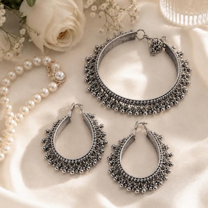 May include: A silver-toned metal jewellery set consisting of a circular choker necklace and two matching teardrop-shaped hoop earrings, each featuring a textured finish and a dangling beaded fringe along the outer edge, accompanied by a pearl necklace.