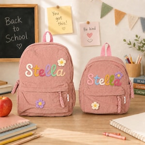 Puede incluir: Dos mochilas de pana rosa con el nombre Stella en letras bordadas de colores. Cada mochila tiene un bolsillo frontal con cremallera, un asa superior y una pequeña flor de ganchillo. La mochila grande tiene una flor blanca, mientras que la pequeña tiene una flor morada en la parte superior y una flor blanca en el bolsillo inferior.