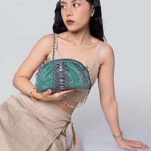 Może przedstawiać: Zielona i czarna kopertówka ze skóry węża ze złotym łańcuszkiem. Torba ma zaokrąglony kształt i teksturowaną powierzchnię. Kopertówka jest trzymana przez osobę.