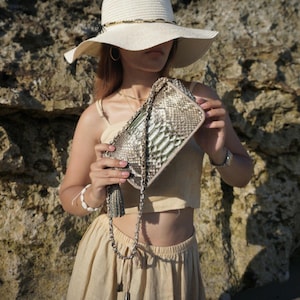 May include: A beige sun hat with a black trim and a snake-skin patterned clutch with a silver chain strap. The clutch has a tassel and is held by a person wearing a beige outfit. The background is a rocky surface.