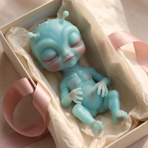 May include: A small, light blue alien figurine with a smooth, glittery finish, large closed eyes, and two antennae on its head. The figure has a rounded belly and delicate limbs, resting inside a rectangular box lined with light-colored tissue paper.