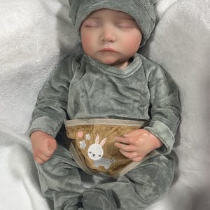 Op de afbeelding: Een realistische babypop gekleed in een grijze pluche outfit en een bijpassende hoed met berenoren. De pop heeft gesloten ogen en een bruine luier met een konijn- en bloemenontwerp.