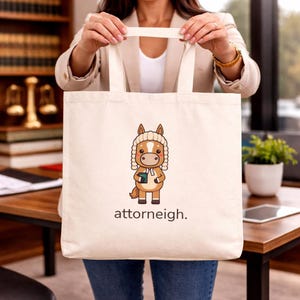 May include: A cream-colored canvas tote bag featuring a cartoon illustration of a brown horse wearing a white barrister wig and a white neck tab. The horse holds a small green book. Below the illustration, the text reads "attorneigh."