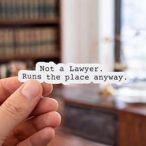 May include: A white die-cut vinyl sticker with a black typewriter-style font. The text reads: Not a Lawyer. Runs the place anyway. The sticker has a smooth, matte finish and a clean, irregular border that follows the shape of the text.