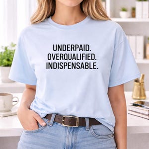 May include: A light blue short-sleeve t-shirt featuring the black text "UNDERPAID. OVERQUALIFIED. INDISPENSABLE." printed in a clean, sans-serif font across the chest. The shirt has a classic crew neckline and a standard fit.