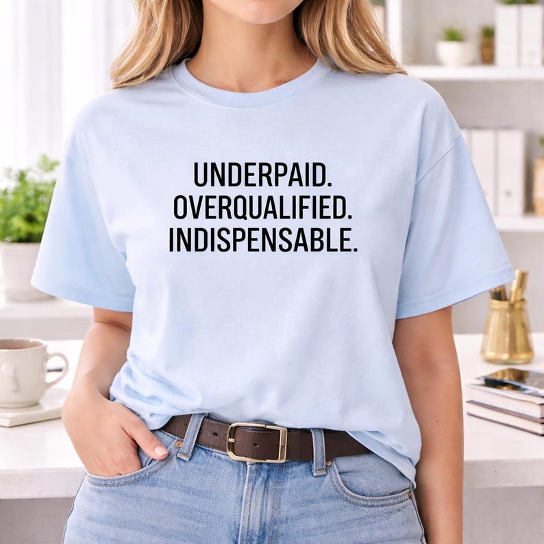 May include: A light blue short-sleeve t-shirt featuring the black text "UNDERPAID. OVERQUALIFIED. INDISPENSABLE." printed in a clean, sans-serif font across the chest. The shirt has a classic crew neckline and a standard fit.