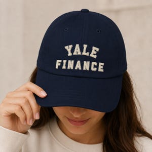 Op de afbeelding: Een marineblauwe baseballpet met de tekst YALE FINANCE in wit borduurwerk, waarbij de letters een gestreept textuurpatroon hebben. De pet heeft een gestructureerde kroon, een gebogen klep en kleine ventilatiegaatjes op de bovenkant.