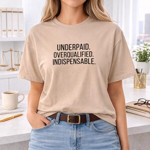 May include: A beige t-shirt with the text "UNDERPAID. OVERQUALIFIED. INDISPENSABLE." in black block letters. The shirt is worn with blue jeans and a brown belt with a gold buckle. The person is standing in front of a white countertop.