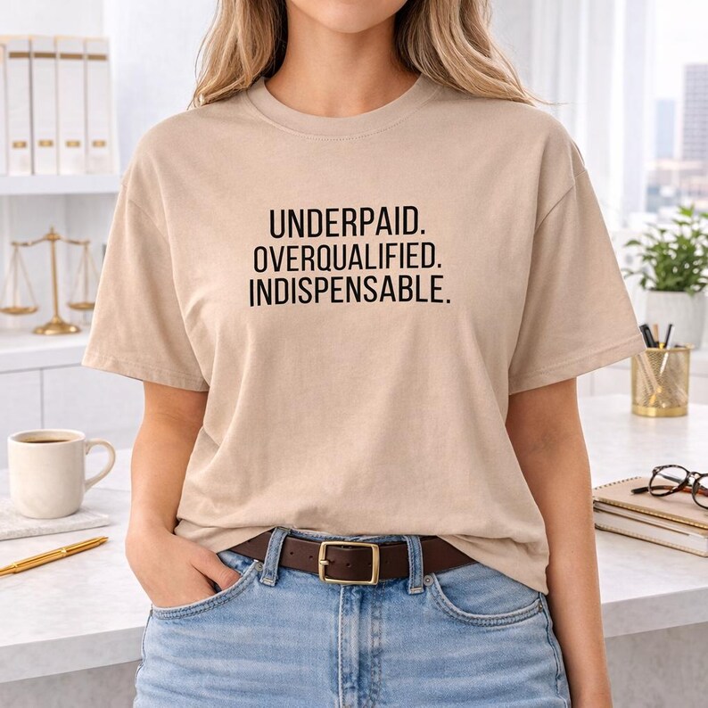 May include: A beige t-shirt with the text "UNDERPAID. OVERQUALIFIED. INDISPENSABLE." in black block letters. The shirt is worn with blue jeans and a brown belt with a gold buckle. The person is standing in front of a white countertop.