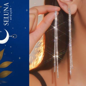 Könnte beinhalten: Lange, silberfarbene Ohrringe mit mehreren Strängen aus funkelnden Strasssteinen. Die Ohrringe sind hängend dargestellt und fangen das Licht ein. Das Bild zeigt auch den Text "SELUNA ATELIER" auf einem dunkelblauen Hintergrund.