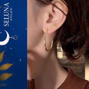 Puede incluir: Pendientes de aro dorados. Los pendientes tienen un diseño sencillo y elegante, con un acabado liso y pulido. Los pendientes se llevan en la oreja de una persona. El fondo es azul oscuro con una luna creciente y el texto "SELUNA - ATELIER".
