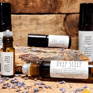 May include: A close-up of a brown glass roller bottle with a black cap and a white label that reads "Deep Sleep 100% Natural". The bottle is surrounded by dried lavender flowers and other botanicals. The label also lists the ingredients, which include chamomile, lavender, vetiver, sweet marjoram, cedarwood, and valerian. The bottle is 3.5 oz/10 ml.