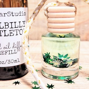 May include: A brown glass bottle with a black dropper top and a white label that reads "A Cellar Studio Hillbilly Mistletoe scented oil diffuser REFILL 1oz/30ml". The bottle is next to a clear glass bottle with a wooden top and a gold string. The bottle is filled with a clear liquid and green glitter. Both bottles are on a wooden surface with green glitter scattered around them.