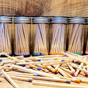 May include: Ten glass jars with silver lids filled with wooden matches. The matches have colored tips in various colors, including blue, green, orange, and pink. The jars are arranged in a row on a wooden surface.