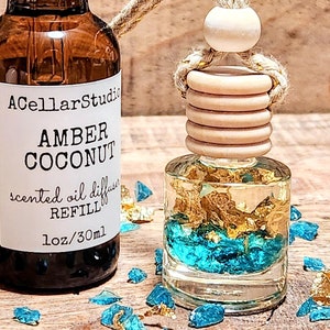 May include: A brown glass bottle with a black dropper top and a white label that reads "ACellarStudio Amber Coconut scented oil diffuser REFILL 1oz/30ml". The bottle is next to a clear glass car air freshener with a wooden top and gold flakes and blue glass pieces inside. The car air freshener is hanging from a brown rope.