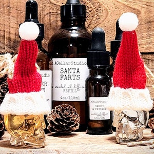 May include: Three glass bottles with gold skull bottle stoppers wearing red and white knit Santa hats. The bottles are filled with a clear liquid and have labels that read "Santa Farts Scented Oil Diffuser Refill 4oz/119ml" and "Sweet & Twisted Scented Oil Diffuser Refill 0.5oz/15ml".