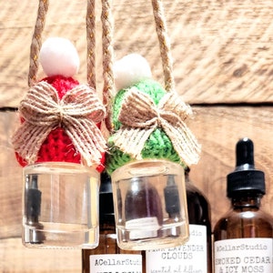 May include: Four glass bottles with droppers filled with scented oil diffuser refills. The bottles are labelled with the scents: "Hillbilly Mistletoe", "Lavender Clouds", "Smoked Cedar & Icy Moss", and "Smoked Cedar & Icy Moss". The bottles are sitting on a wooden surface. Two small glass bottles with droppers are hanging from crocheted hats.