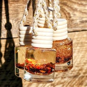 May include: Three small, square glass bottles filled with a shimmering, amber-colored liquid and glitter. Each bottle has a light wood cap and is suspended by a braided, cream-colored cord with gold accents. The background is a wooden surface.