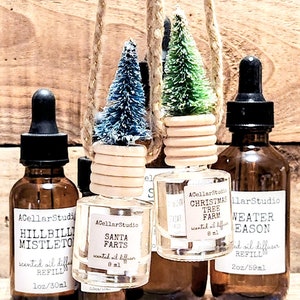 以下が含まれることがあります： 茶色の蓋とラベルが付いたガラス瓶が4本。瓶には香りのオイルディフューザーが入っています。ラベルには「ヒルビリー・ミストletoe」、「サンタ・ファーツ」、「クリスマスツリーファーム」、「ウィンターシーズン」と書いてあります。瓶は木製の表面に置かれ、その上に2本の小さな人工の松の木がぶら下がっています。