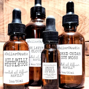May include: Four amber glass bottles with dropper tops and labels. The labels are white with black text. The bottles contain scented oil diffuser refills. The scents are Hillbilly Mistletoe, Smoked Cedar & Icy Moss, Pink Lavender Cloud, and Smoked Cedar & Icy Moss. The bottles are 1 oz/30 ml.
