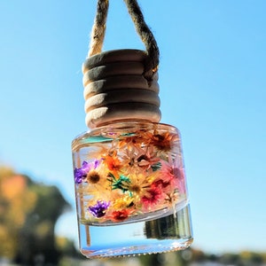 Puede incluir: Una botella de vidrio transparente con una tapa de madera cuelga de una cuerda marrón. La botella está llena de flores secas en tonos de rosa, amarillo, naranja, morado y verde.