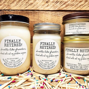 K&ouml;nnte beinhalten: Drei Glasbeh&auml;lter mit Sojawachskerzen. Jedes Glas hat ein wei&szlig;es Etikett mit dem Text "FINALLY RETIRED! Smells like freedom, infused with not my problem anymore." Die Gl&auml;ser stehen auf einer Holzoberfl&auml;che, umgeben von bunten Streichh&ouml;lzern.