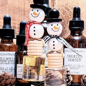 May include: A set of four glass bottles with snowman-shaped bottle stoppers. The bottles are filled with a clear liquid and have black droppers. The bottles are labeled with the text "CellarStudio", "Hillbilly Mistletoe", "Sweater Season", and "scented oil diffuser refill 2/59ml".