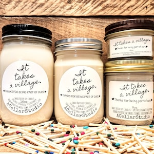 May include: Three glass jars with brown, gold, and black lids. Each jar contains a soy candle with a white label that reads "It takes a village. Thanks for being part of ours." The labels also include the company name "ACellarStudio" and the size of the candle in ounces. The jars are sitting on a surface covered in colorful matchsticks.
