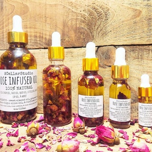 May include: Four glass bottles of rose infused oil in various sizes with gold lids and droppers. The bottles are filled with rose petals and a clear oil. The bottles are sitting on a wooden surface with rose petals scattered around them. The labels on the bottles read "Rose Infused Oil 100% Natural".
