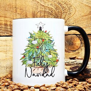 Puede incluir: Taza de cerámica blanca con asa negra. La taza presenta un diseño de árbol de Navidad con hojas verdes, adornos rojos y blancos y una estrella en la parte superior. El texto "Feliz Navidad" está escrito debajo del árbol.