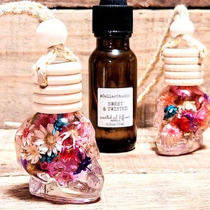 May include: Two glass skull car air fresheners with dried flowers inside. The fresheners are hanging from a white string. A small bottle of essential oil diffuser refill is in the background. The bottle label reads "A Collar Studio Sweet & Twisted Scented Oil Diffuser Refill 0.5oz/15ml."
