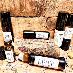 May include: Five brown glass roller bottles with black caps and labels that say "Deep Sleep 100% Natural". The bottles are arranged on a wooden surface with dried lavender flowers.