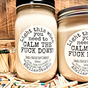 May include: Two soy candles in glass jars with black lids. The labels read "Light this when you need to CALM THE FUCK DOWN!" and "Small Batch Soy Candles." Matches with colored tips are scattered around the candles.