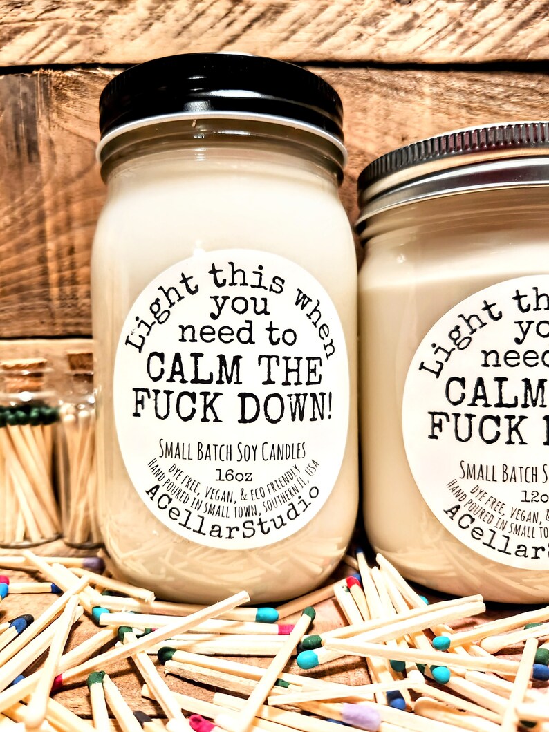 May include: Two soy candles in glass jars with black lids. The labels read "Light this when you need to CALM THE FUCK DOWN!" and "Small Batch Soy Candles." Matches with colored tips are scattered around the candles.