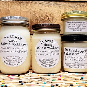 May include: Three glass jars with white soy candles. Each jar has a white label with the text "It truly does take a village, & we are so grateful you are part of ours!" and the brand name "ACellarStudio". The jars are arranged in a pyramid shape on a wooden surface.